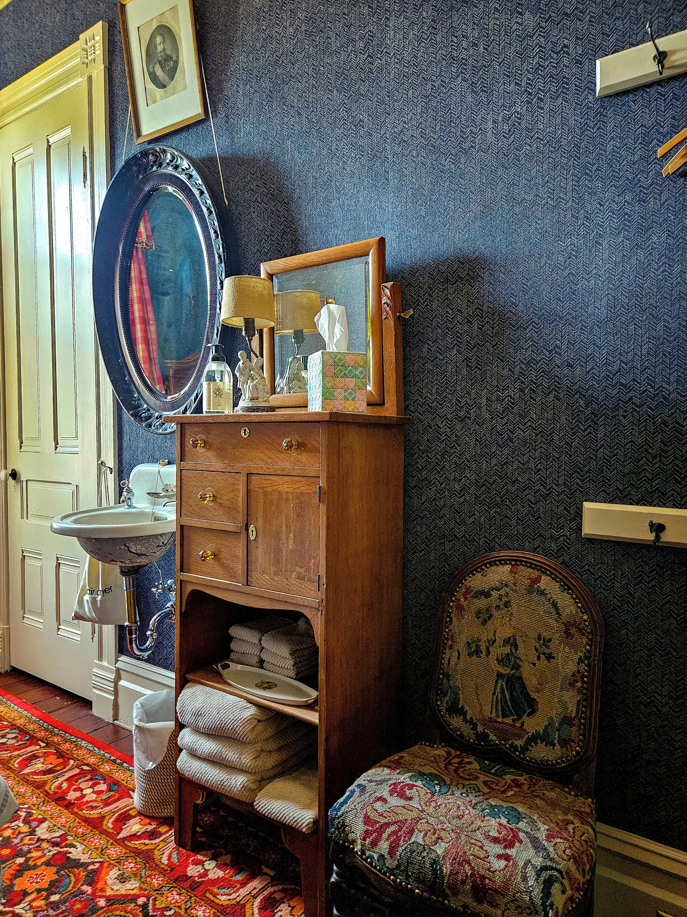 Quarto antigo com cômoda de madeira, um espelho, uma cadeira com almofada estampada e um tapete colorido.