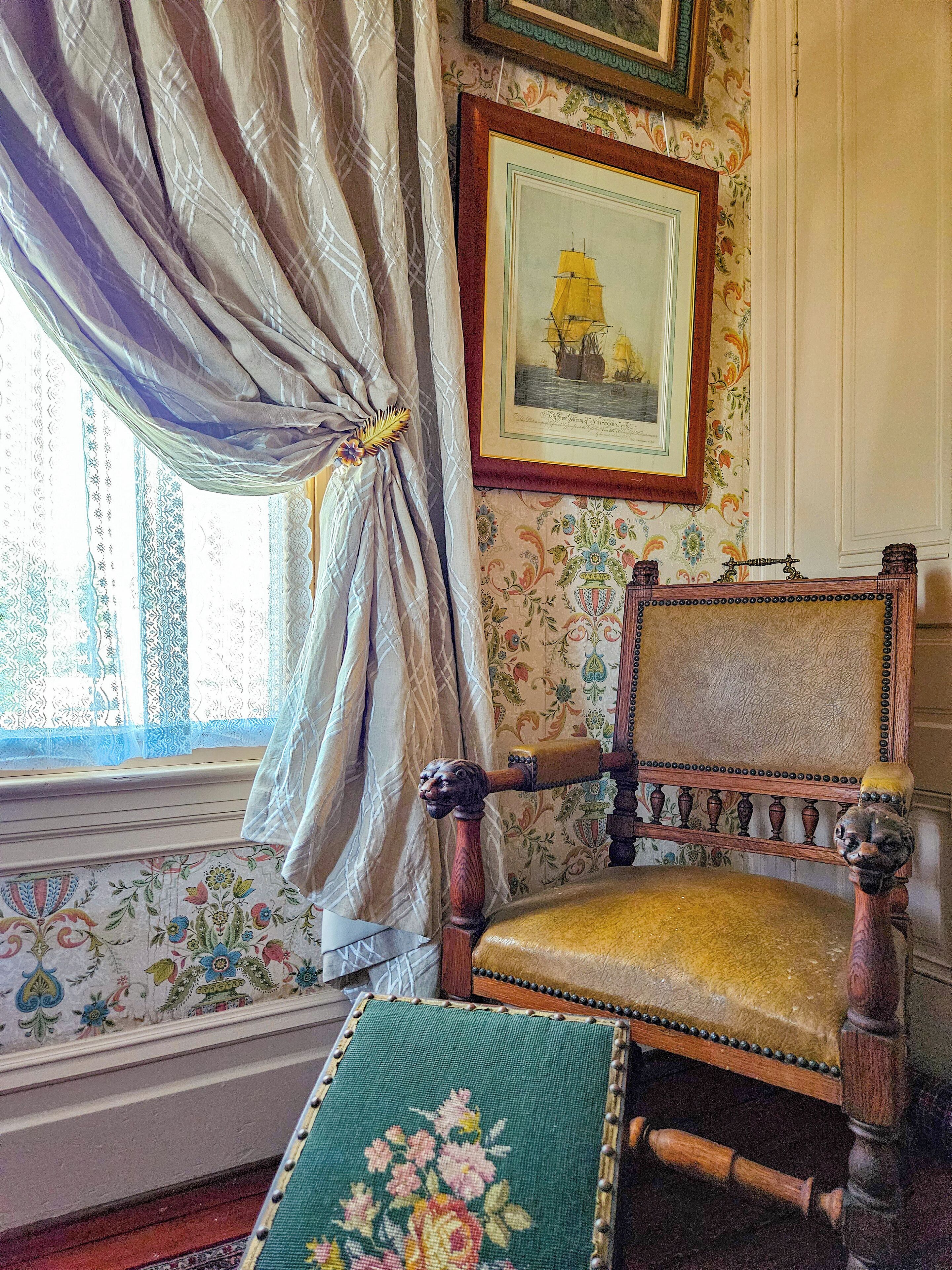 A vintage chair with a floral cushion, a framed picture of a ship, and patterned wallpaper.
