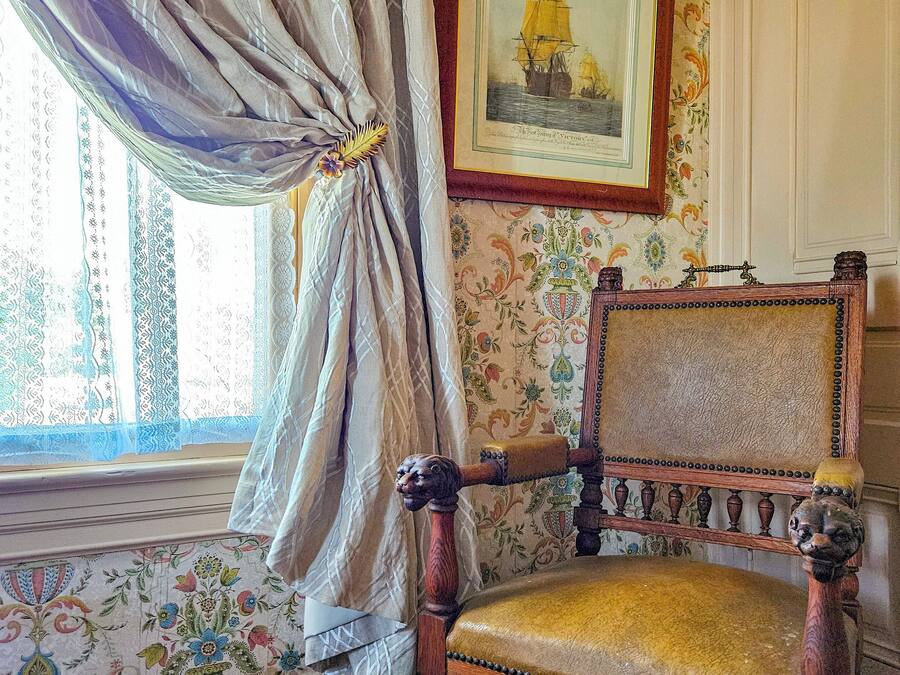 A vintage chair with a floral cushion, a framed picture of a ship, and patterned wallpaper.