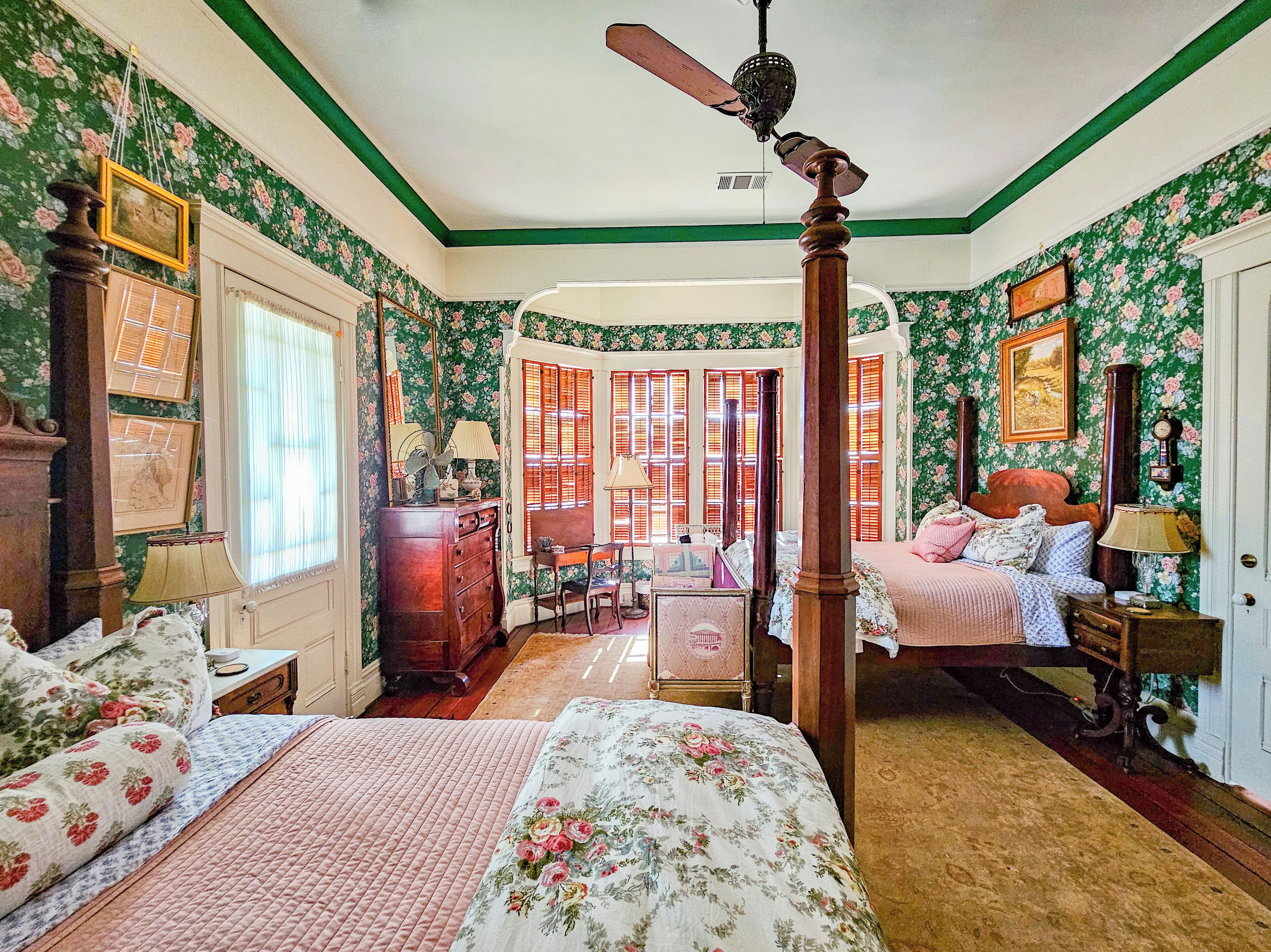 A room with a four-poster bed, a wooden desk, and a chair. There are two windows with shutters, a ceiling fan, and a door.