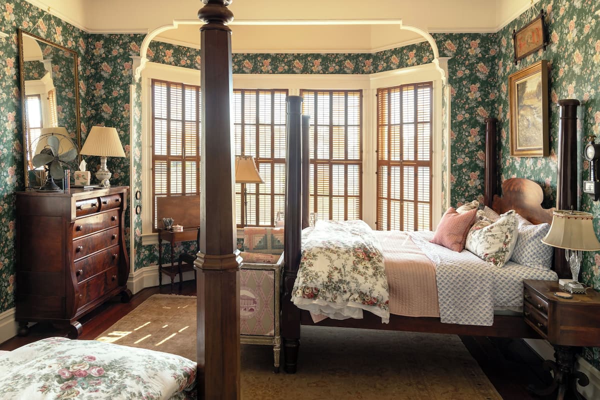 A bedroom with a four-poster bed, floral wallpaper, and a bay window with shutters.