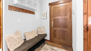 A hallway with a wooden door, a bench with fluffy cushions, a white wall with a coat rack, and a framed picture. - Cv112c-2 (Copper Mountain)