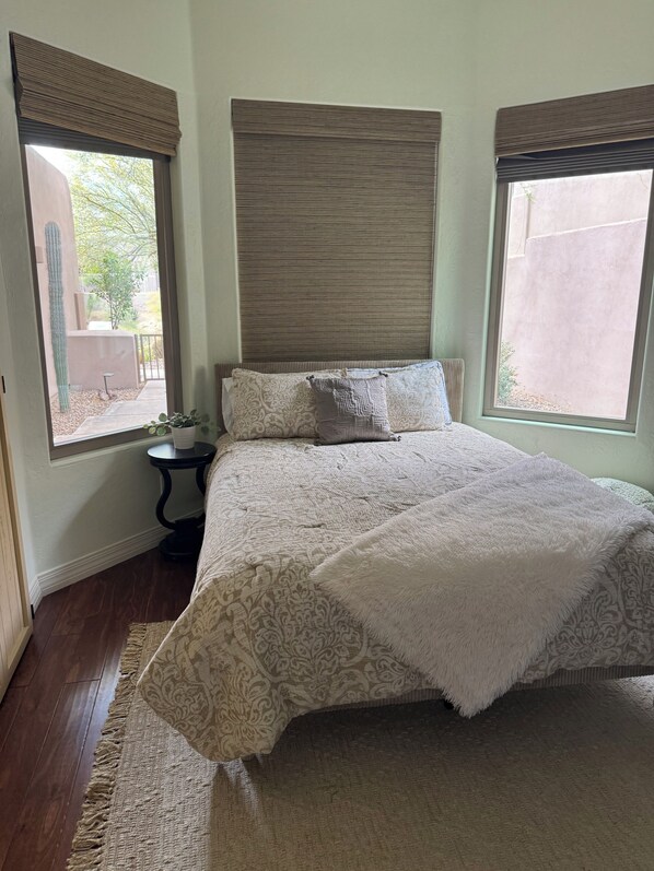 A bedroom with a large bed, a bedside table, a window with a view of a cactus and trees, and a headboard with vertical lines.