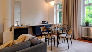 Un soggiorno moderno con divano, tavolo da pranzo con sedie, angolo cottura con piano di lavoro ed elettrodomestici, e un'ampia finestra con tende.