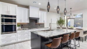A modern kitchen with white cabinets, a large island with bar stools, a built-in oven, and pendant lighting.