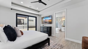 A modern bedroom with a large bed, a ceiling fan, a sliding glass door leading to a balcony, and a painting on the wall.