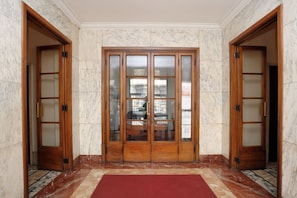 A hallway with marble walls and a red carpet leading to a set of wooden double doors with glass panels.
