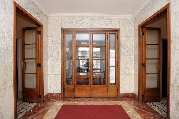 A hallway with marble walls and a red carpet leading to a set of wooden double doors with glass panels.