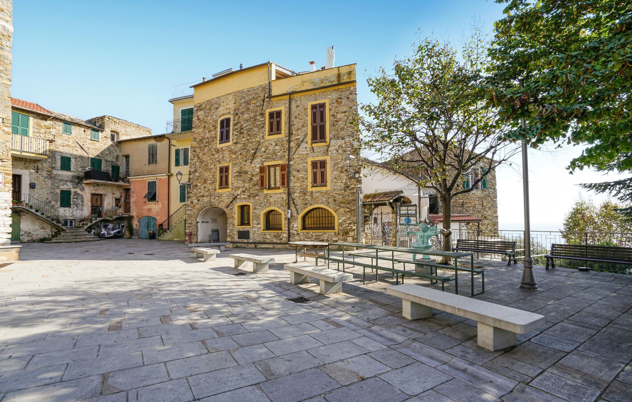 A stone building with multiple windows and a balcony, a paved square with benches, and a tree.