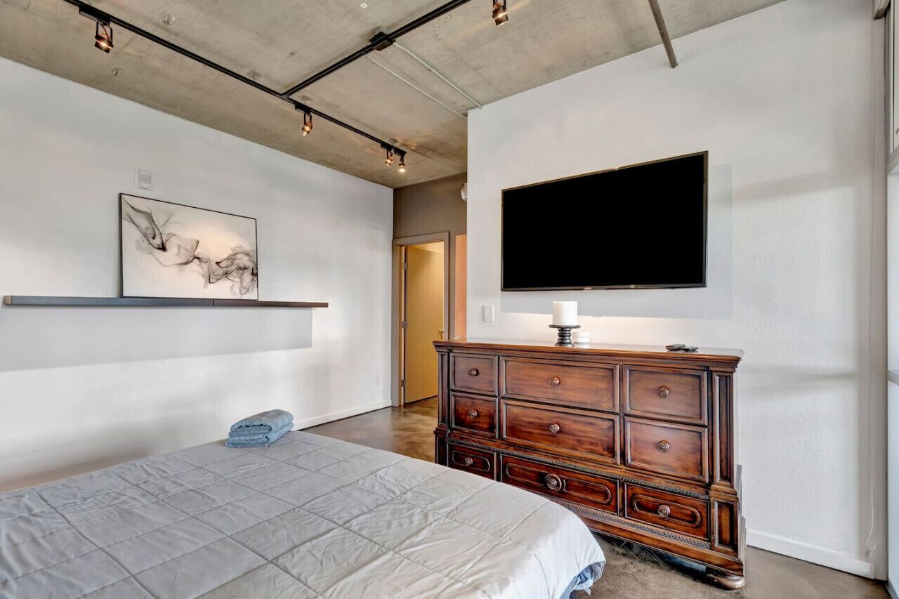 A bedroom with a bed, a dresser, a mounted TV, and a painting on the wall.