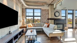 A modern living room with a large flat-screen TV, a sectional sofa, a fireplace, and large windows offering a city view. - Luxurious Uptown Denver Loft with Stunning Views (Denver)