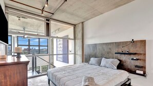 A modern bedroom with a large bed, a wooden dresser, and a view of the city through large windows. - Luxurious Uptown Denver Loft with Stunning Views (Denver)