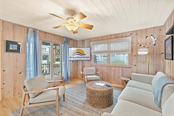 A cozy living room with wooden walls, a ceiling fan, a large TV, and a wicker ottoman.
