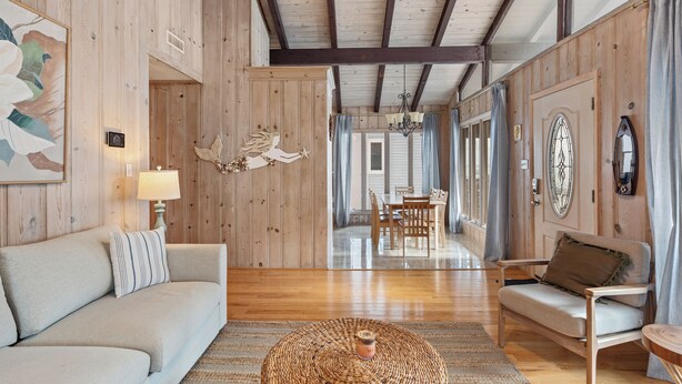 A spacious room with wooden walls and ceiling, a sofa, a chair, a dining table, and a large window with curtains.