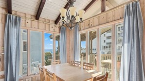 A wooden dining area with a table and chairs, large windows with curtains, and a chandelier.