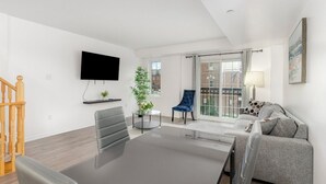 Un salón moderno con una mesa de comedor, un sofá, un televisor en la pared y un balcón con vistas a edificios.