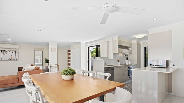 A modern kitchen with white cabinets, a dining area with a wooden table and white chairs, and a ceiling fan. - Relax at Riviera (Miami)