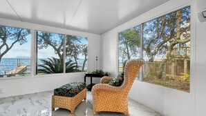 A spacious room with large windows, a wicker chair, a matching ottoman, and a small table with a plant. - Baywatch Waterfront Paradice  8 m to bch, private pier, Sleeps 8 Fenced Dog Area (Gulf Shores)