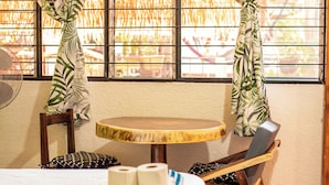 A room with a round wooden table, two wooden chairs, a bed with a white sheet, and a window with metal bars and curtains with a leaf pattern. - Casa San Jeronimo (San Pedro Mixtepec)