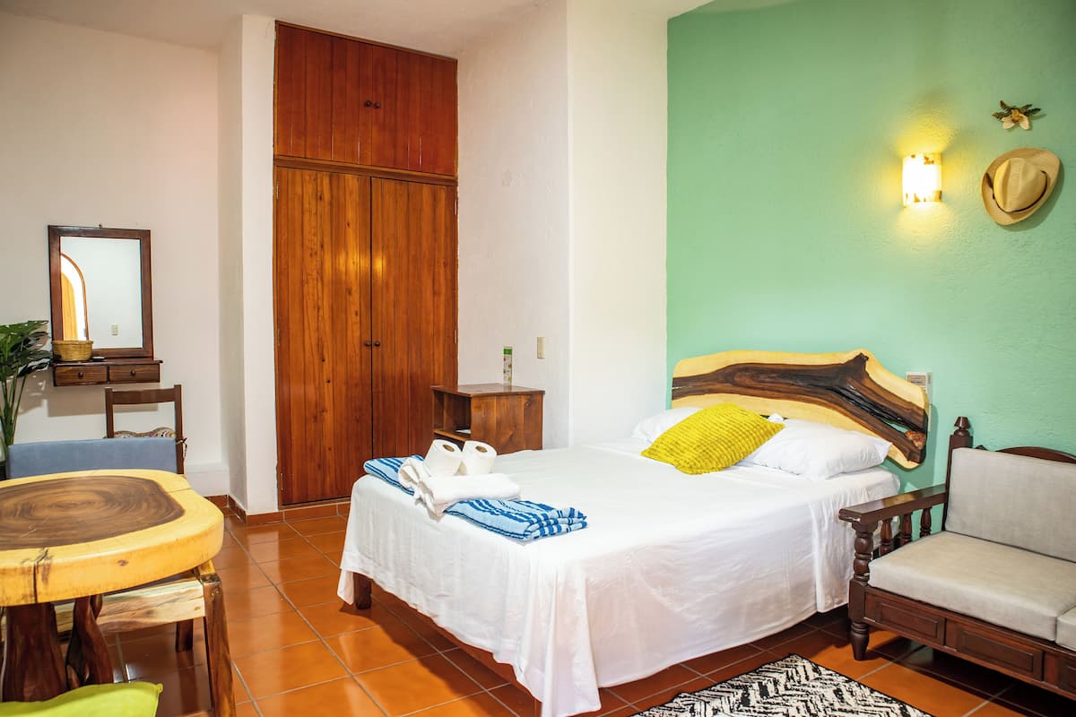 A hotel room with a bed, a wooden wardrobe, a small table with chairs, and a wall-mounted light fixture.