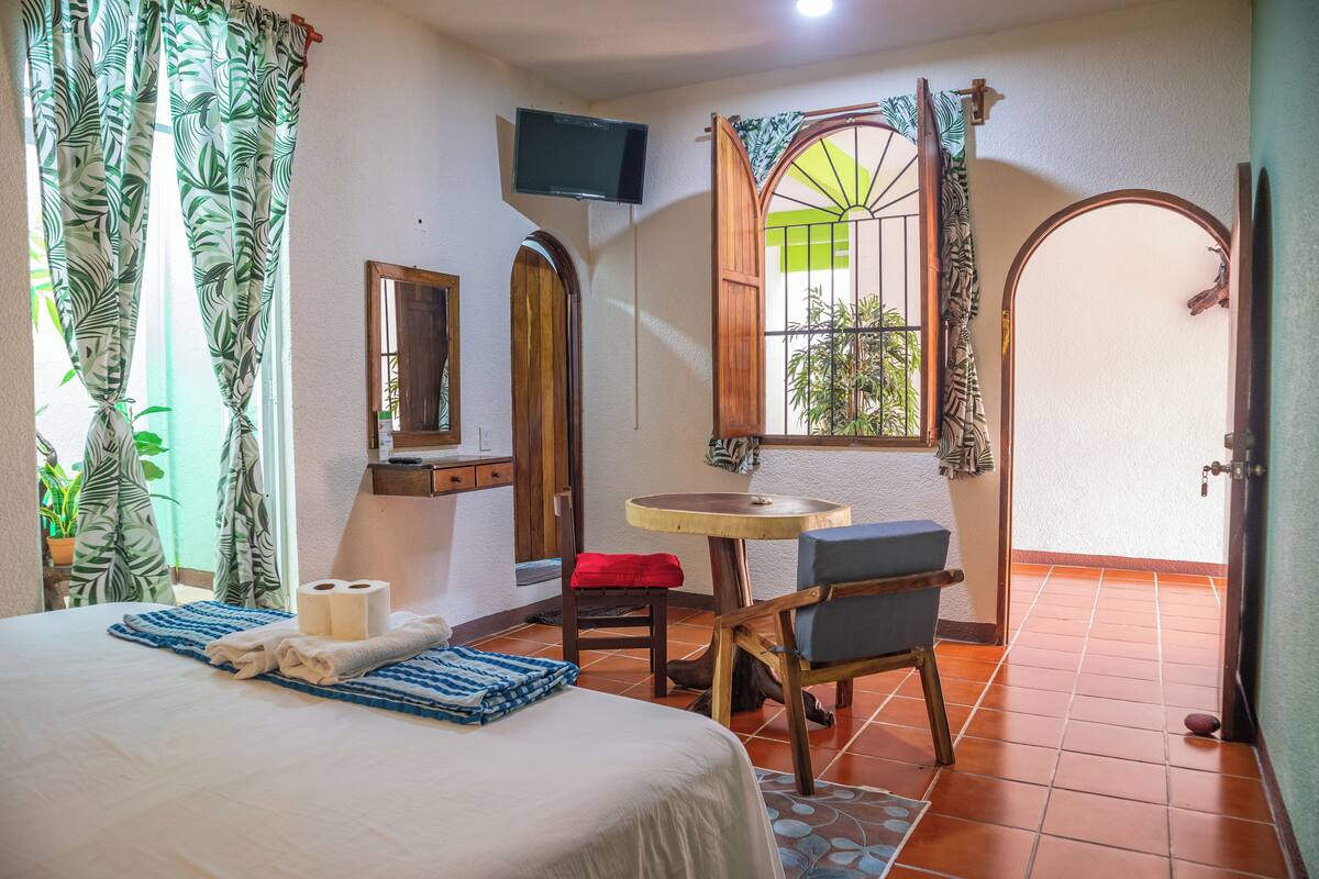 A hotel room with a bed, a small table with a chair, a TV mounted on the wall, and a window with curtains.