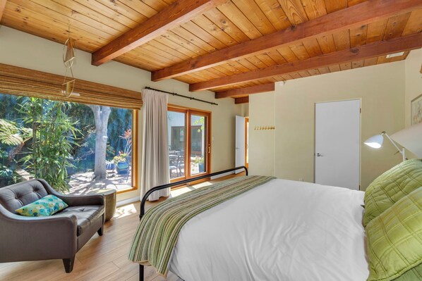 Un dormitorio con cama, una silla, un ventanal con cortinas, una puerta y vistas al exterior.