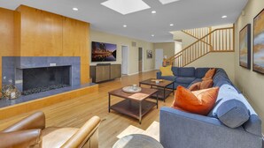 A spacious living room with a fireplace, wooden flooring, a blue sofa, and a staircase. - Sea Breeze Dream | Water View, Rooftop Deck, Game Room, Fire Pit & Cozy Backyard! (San Diego)