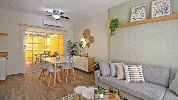 Een moderne woonkamer met een grijze bank, een eettafel en stoelen. Er is een plafondventilator, een plant en kunst aan de muur.
