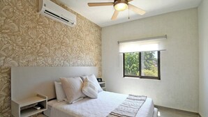 Een slaapkamer met een bed, een plafondventilator, een airconditioningapparaat, een raam met zonwering en een wand met een bloemenmotief.