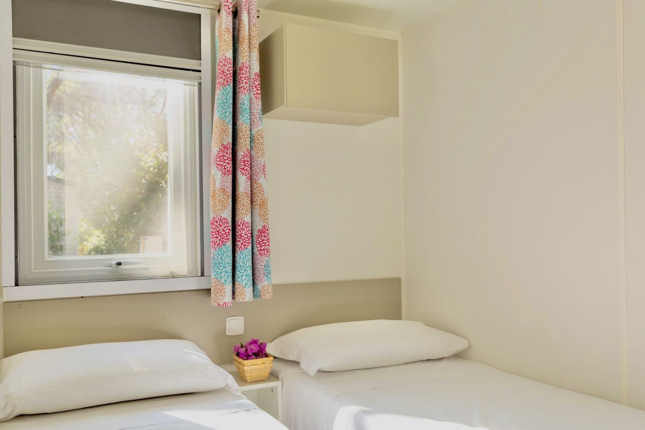 Une chambre d’hôtel avec deux lits, une fenêtre avec des rideaux, une petite table de chevet sur laquelle se trouve un panier de fleurs, et un mur de couleur claire.