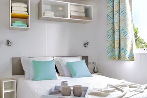 Un dormitorio ordenado con cama, mesitas de noche, un estante con toallas dobladas y una ventana con cortinas estampadas.