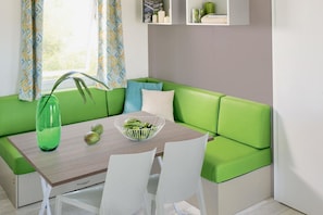 Un espacio habitable moderno con un sofá verde, una mesa de madera y sillas blancas. Un jarrón con flores verdes y un frutero están sobre la mesa.