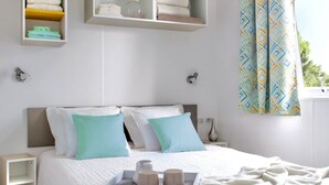 A neatly arranged bedroom with a bed, bedside tables, a shelf with folded towels, and a window with patterned curtains.