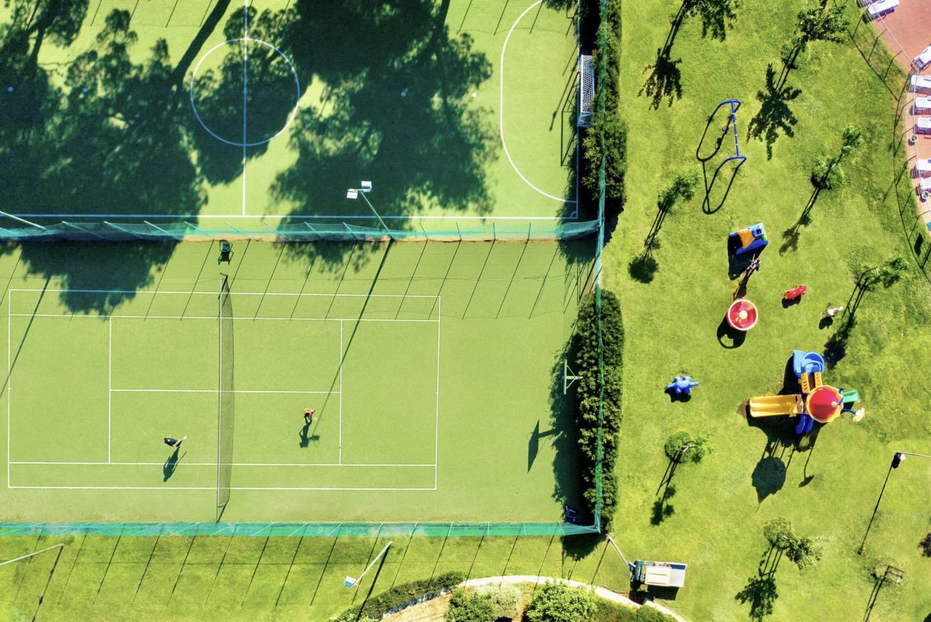 Eine Vogelperspektive auf einen Tennisplatz, umgeben von Bäumen und Spielgeräten.