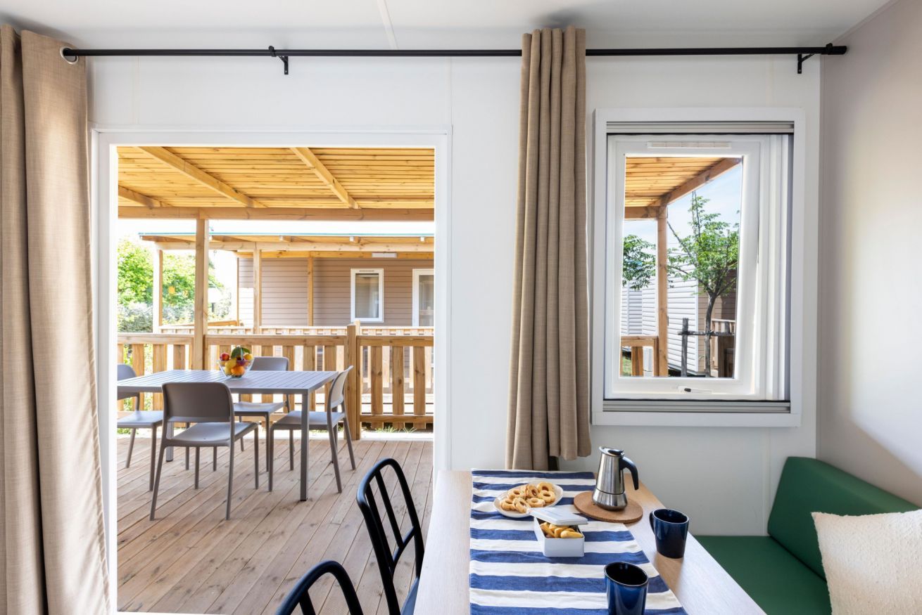 Une pièce comprenant un coin repas, une fenêtre avec des rideaux et donnant sur une terrasse extérieure.