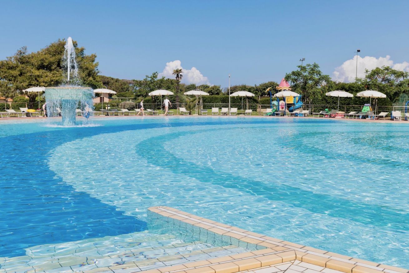 Una piscina con fuente, sillas de descanso y sombrillas.