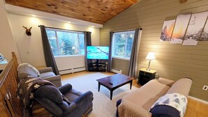 A cozy living room with a wooden ceiling, a television, a sofa, a chair, and a table. - Valley Home with Game Room, Walking Distance to Narrow Gauge Trails & Restaurant (Carrabassett Valley)