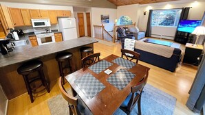 A modern kitchen with wooden cabinets, a microwave, and a refrigerator. There is a dining area with a wooden table and chairs, and a living room with a sofa and a TV. - Valley Home with Game Room, Walking Distance to Narrow Gauge Trails & Restaurant (Carrabassett Valley)