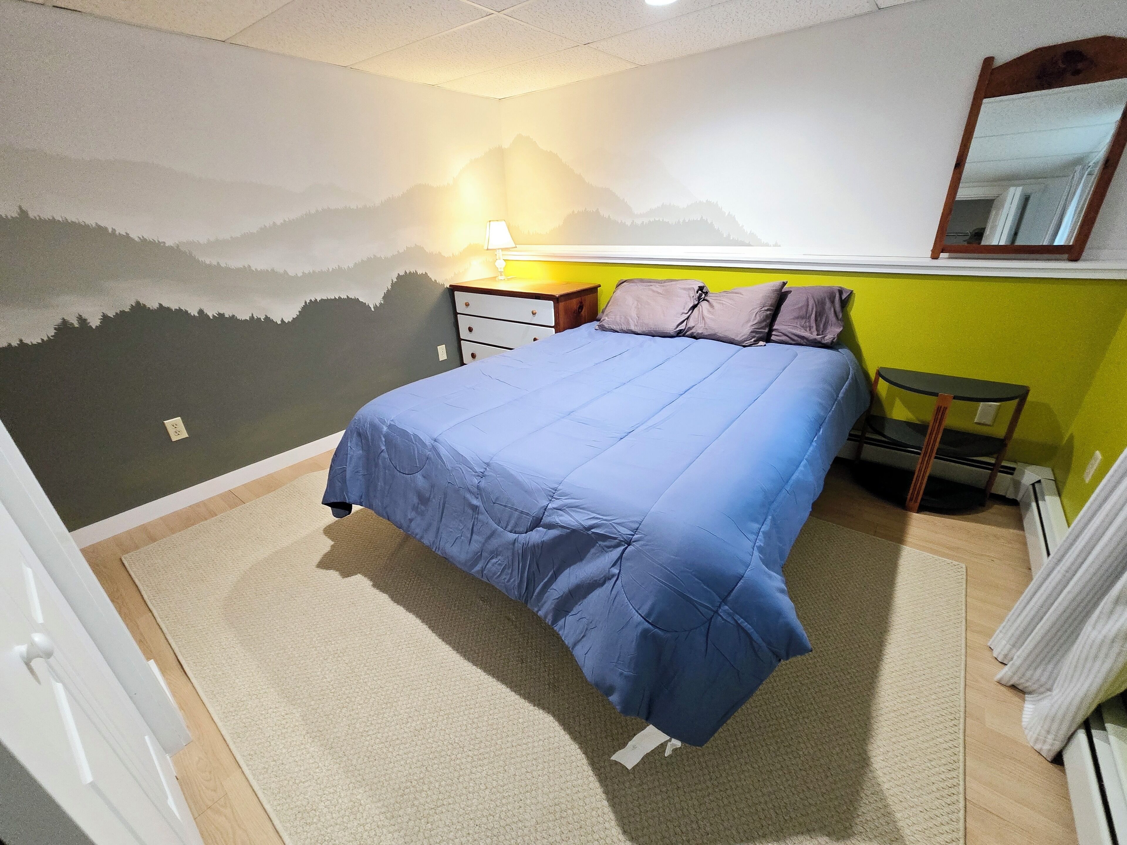 Un dormitorio con cama, mesita de noche, lámpara, silla y un mural en la pared.