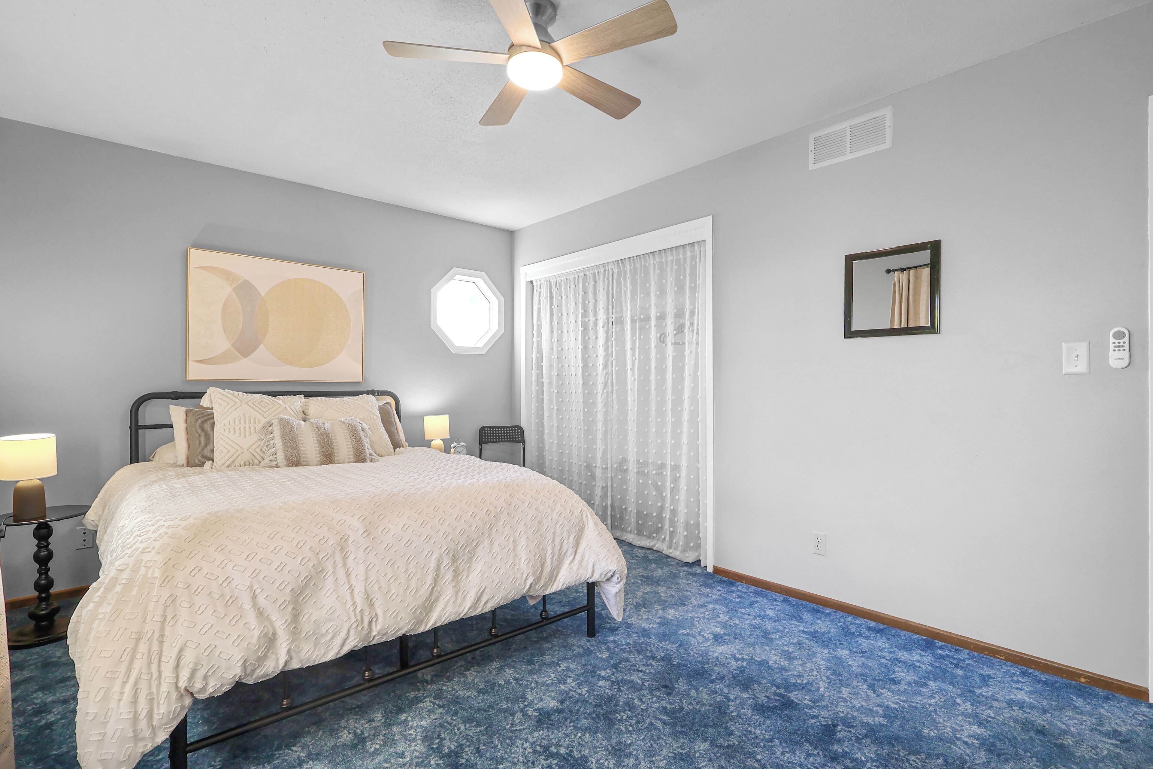 A bedroom with a bed, a ceiling fan, a window with curtains, and a wall-mounted light fixture.