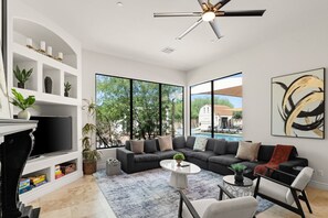 A modern living room with a sectional sofa, a flat-screen TV, a ceiling fan, and a large window overlooking a pool and greenery.