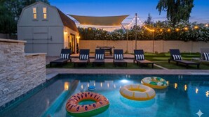 Una piscina con sillas de descanso y flotadores, un espacio de descanso techado y una casa al fondo.