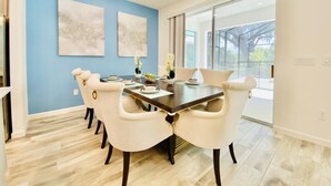 A dining area with a dark wooden table and white upholstered chairs, blue walls, and a large window providing a view of the outdoors. - Amazing 8Br /Private Pool  & hot tub 2547 (kissimmee)