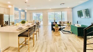 A modern kitchen with a central island, bar stools, and a dining table. There is a living area with a sofa and a coffee table. The room has large windows and a staircase. - Amazing 8Br /Private Pool  & hot tub 2547 (kissimmee)