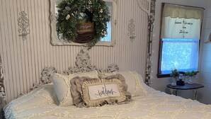 A bedroom with a large bed, a decorative headboard, a wall-mounted mirror, a window with curtains, and a small table with two potted plants. - Charming cottage in Girard with WiFi and AC - perfect getaway (Girard)