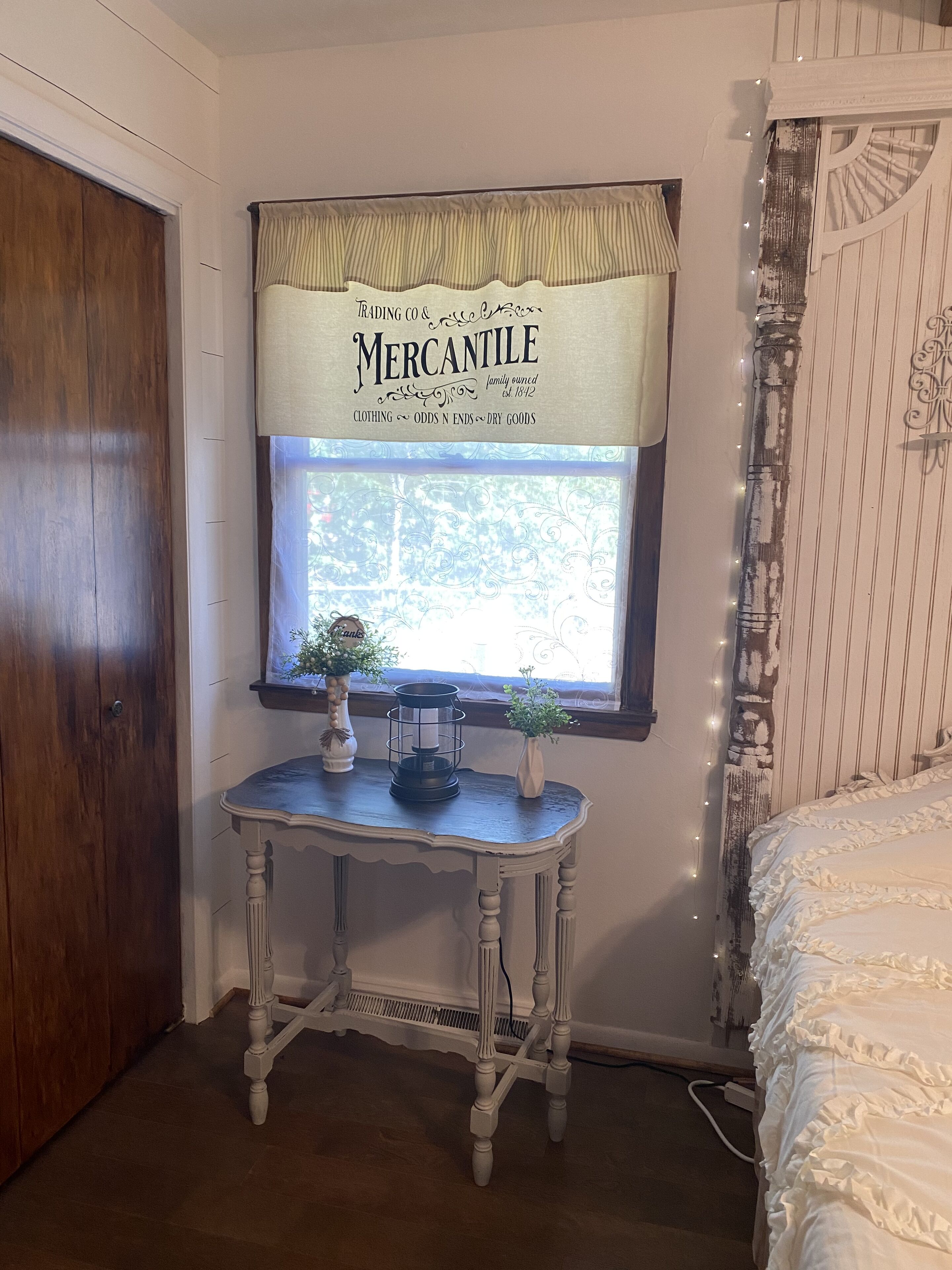 A room with a wooden door, a window with a decorative sign, a small table with a candle holder and a plant, and a bed with ruffled bedding.
