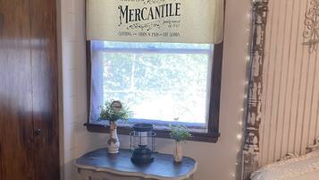 A room with a wooden door, a window with a decorative sign, a small table with a candle holder and a plant, and a bed with ruffled bedding.