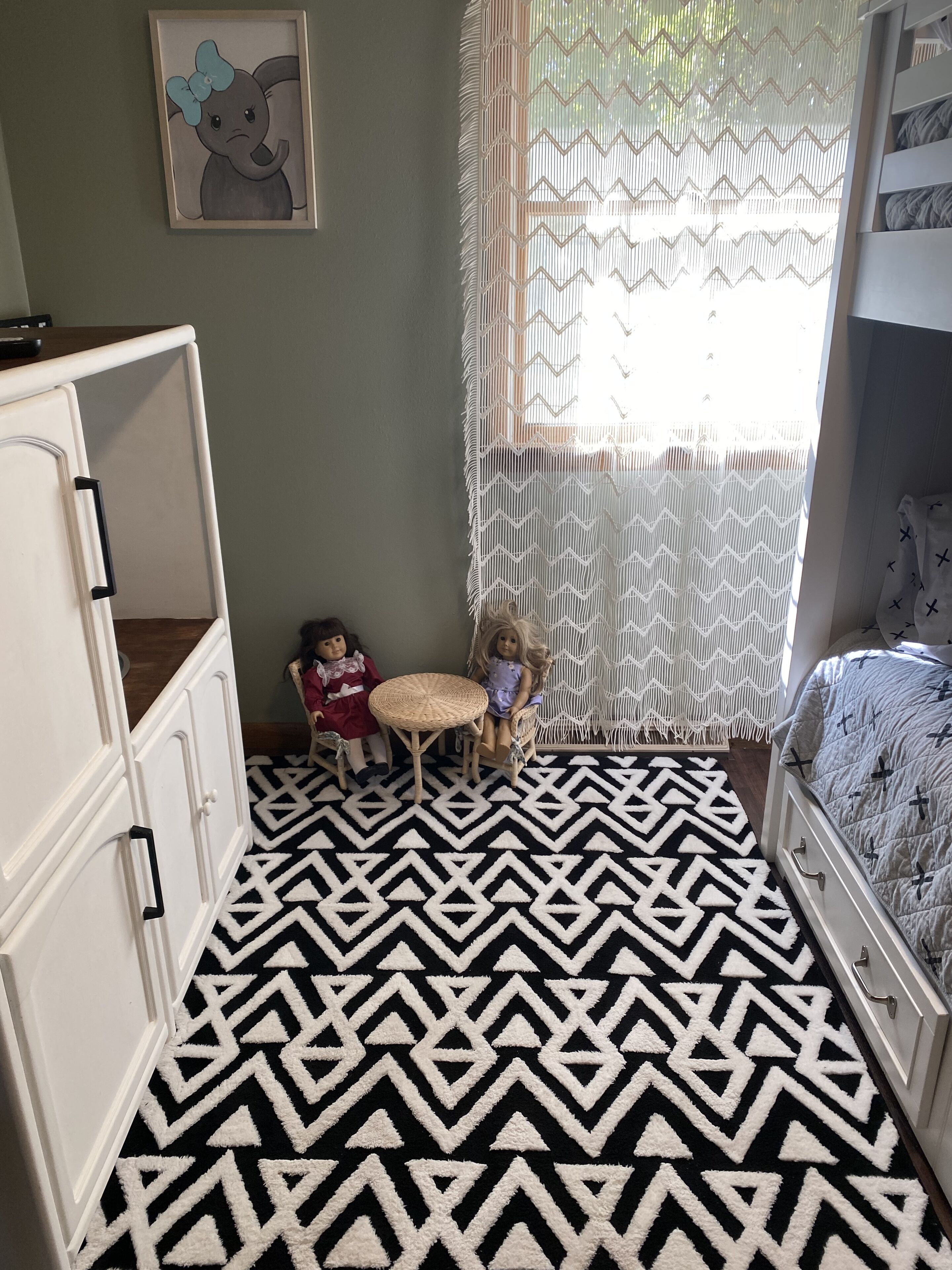 Een kamer met een kamerbreed tapijt met een geometrisch patroon, een wiegje, een klein tafeltje met poppen en een raam met een ruitvormig gordijn.