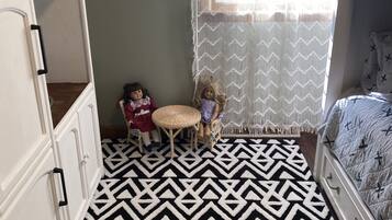 Een kamer met een kamerbreed tapijt met een geometrisch patroon, een wiegje, een klein tafeltje met poppen en een raam met een ruitvormig gordijn.