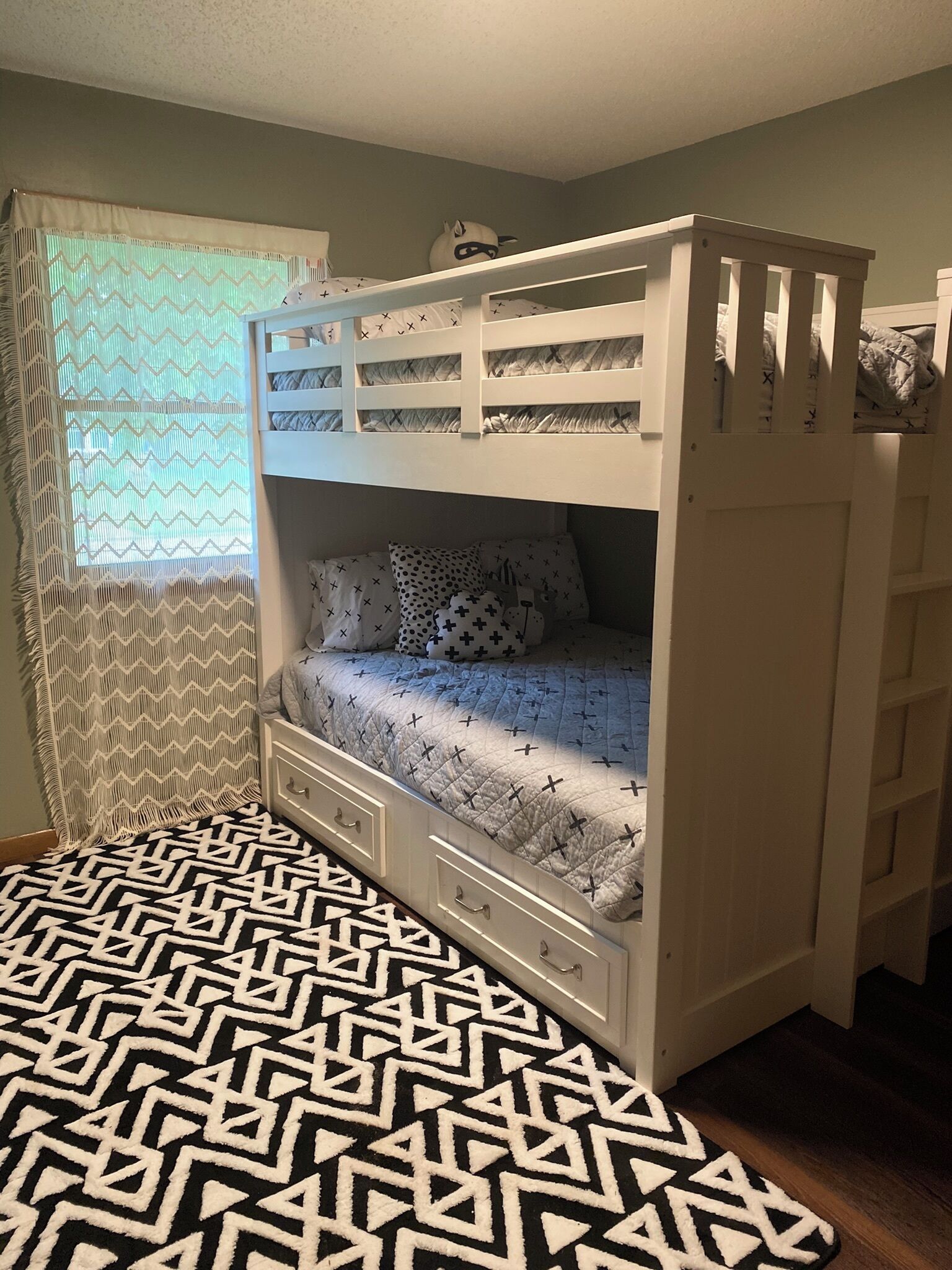 A bunk bed with a patterned bedspread, a window with a zigzag curtain, and a wooden floor.